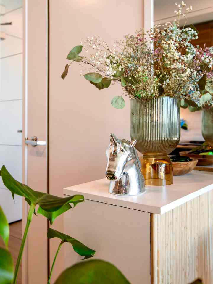 Minimal interior featuring a white table with silver unicorn decoration and dried flowers, accented by green plant leaves in the foreground