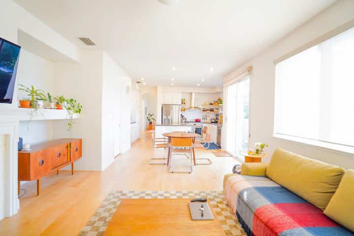 Bright white-toned living room and kitchen featuring wood tone sideboard, mid-century modern dining set and sofa in a minimal interior