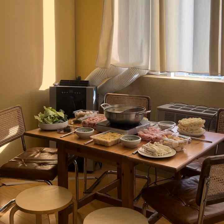 Warm yellow-walled dining room featuring natural wood tone table and chairs, centered hotpot with assorted ingredients for cozy autumn vibe