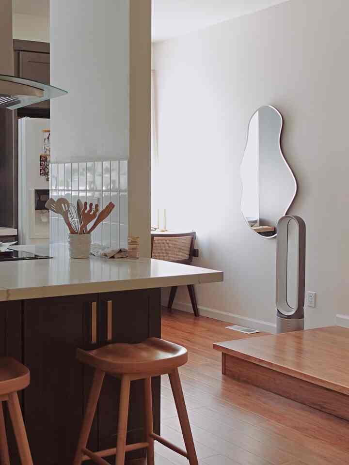 Natural wood tone and white kitchen space featuring a uniquely shaped mirror and wooden bar stools with a cozy atmosphere