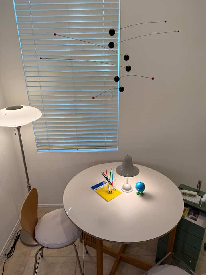 A small mid-century modern workspace featuring a white round table, wood-tone chair, and ambient lighting with blinds