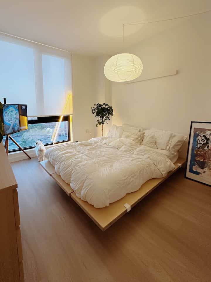 Japandi style bedroom in white and wood tones featuring a large bed, TV stand, and a small dog near the window, creating a cozy atmosphere
