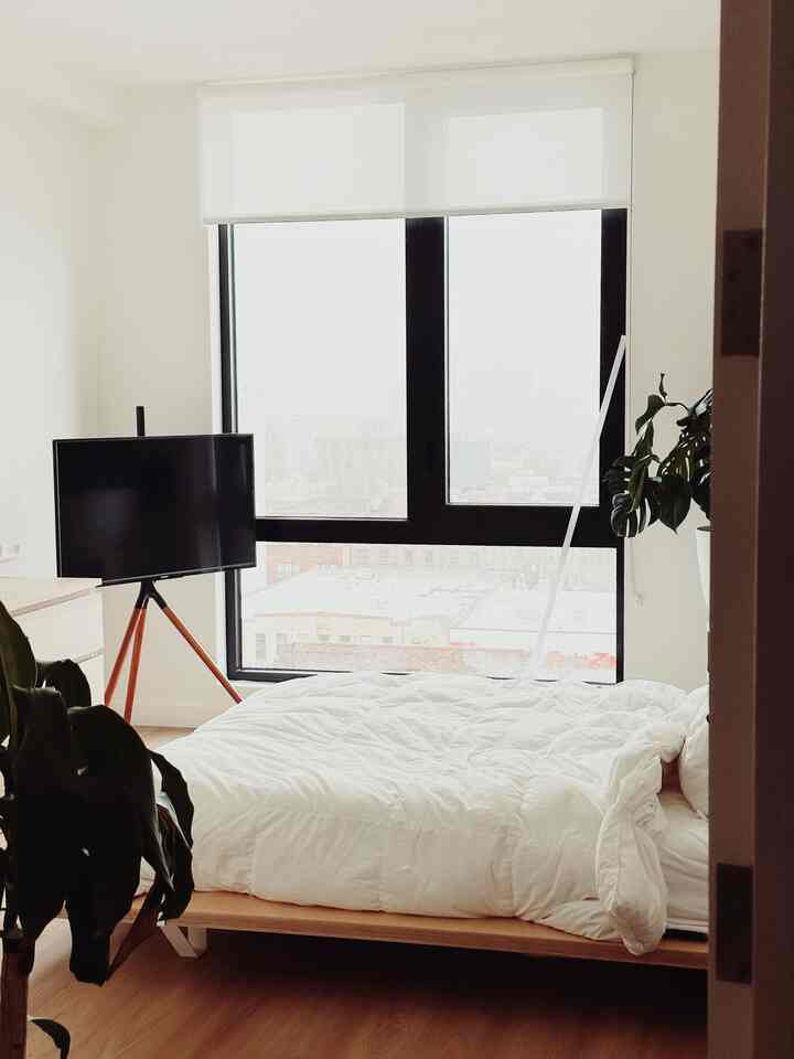 Simple bedroom featuring white tones and wood accents, essentials furniture, and a large window creating a bright atmosphere