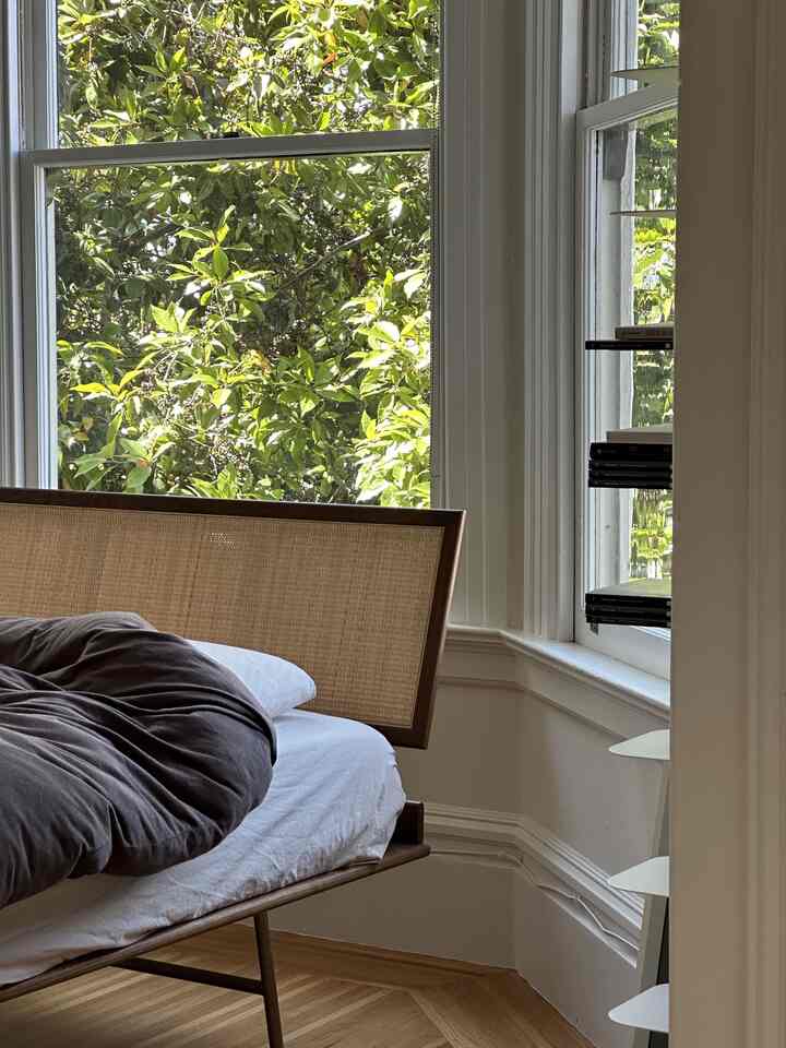 A natural-tone mid-century modern bedroom featuring a wooden bed frame and large windows with lush greenery, creating a cozy atmosphere