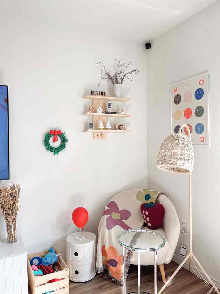 Natural toned living room featuring a cozy armchair, rattan floor lamp, toy storage crate, and small wall shelf