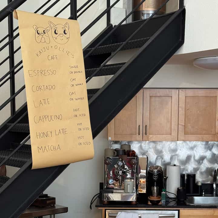 Bright white walls and black metal stairs frame a compact wood-tone kitchen featuring coffee equipment in a home cafe setting