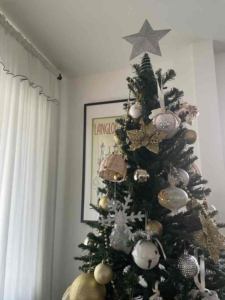 Bright space with white walls and curtains featuring a centrally positioned Christmas tree adorned with gold and silver ornaments, creating a warm festive atmosphere