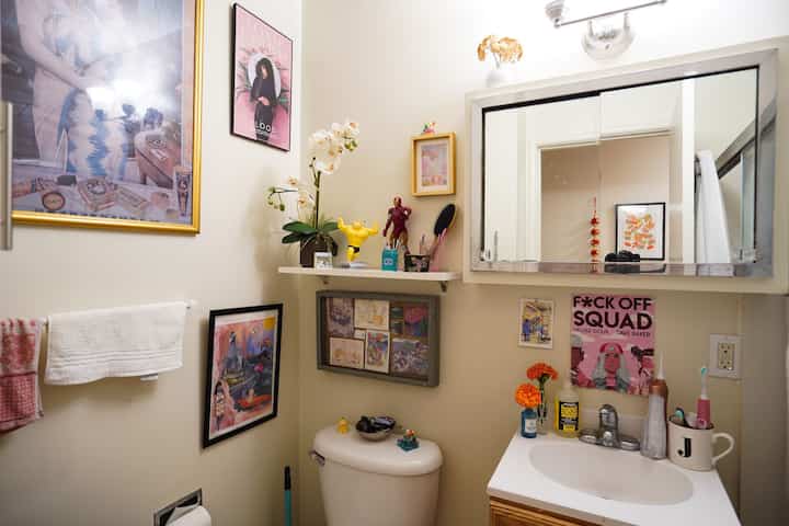 White-toned bathroom featuring a sink, toilet, wall shelf with eclectic decor and framed art, creating a cozy eclectic space
