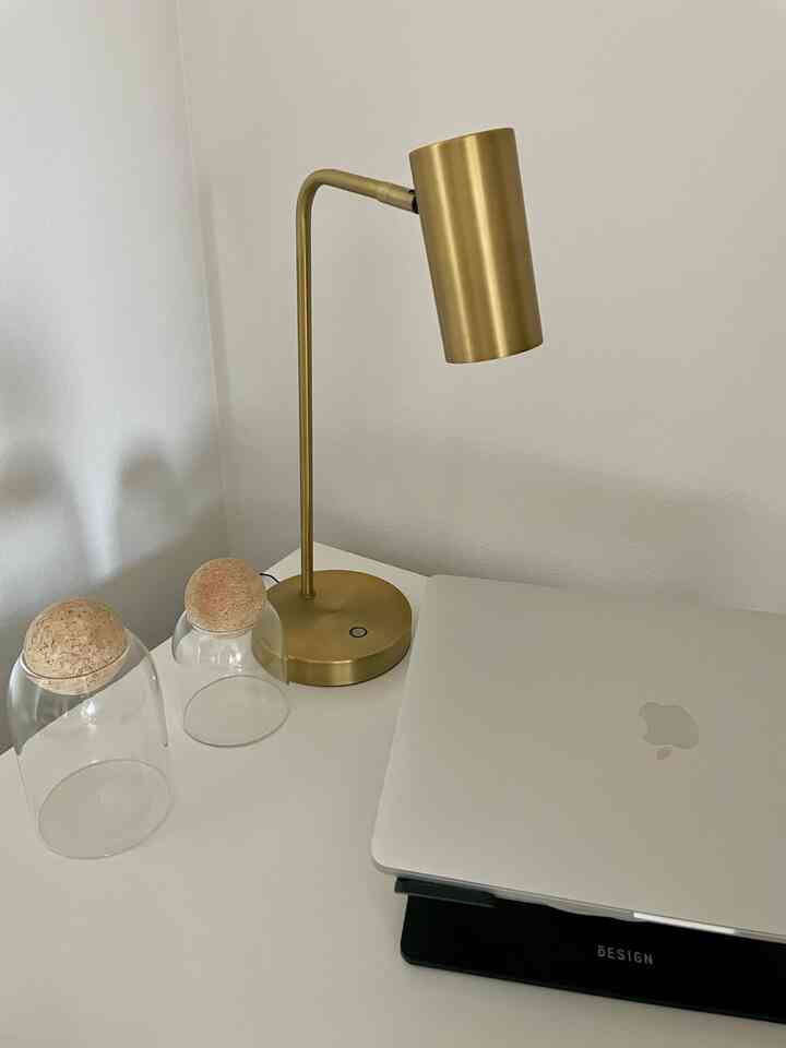A compact home office space with white walls and desk, featuring a silver laptop and gold table lamp on a clean workspace