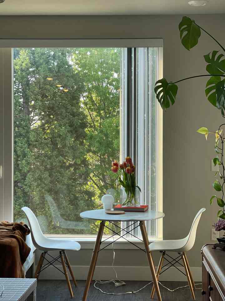 A simple and modern two-person dining room featuring a white round table with wood-toned legs and two white chairs by a large window with natural light