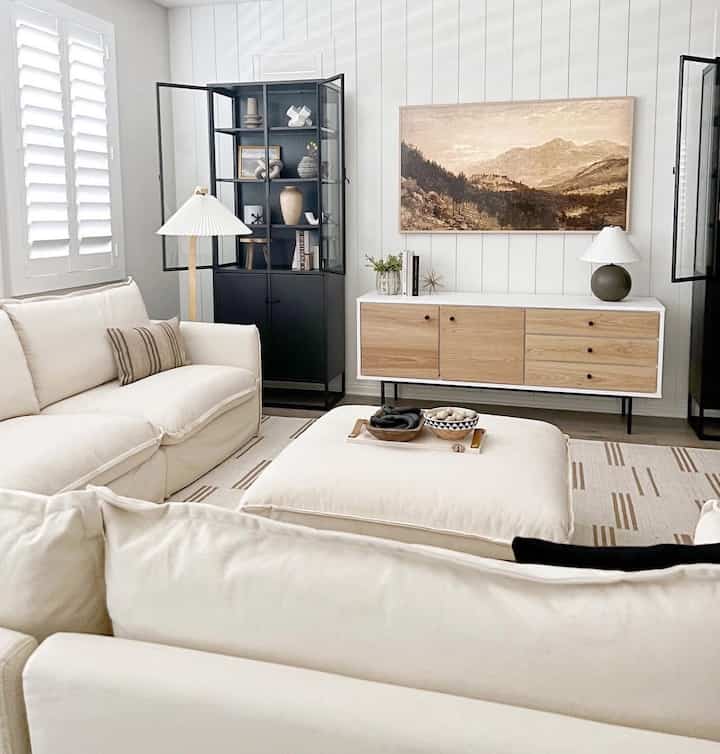A modern living room with white and natural tones featuring a sectional sofa, sideboard, and corner storage cabinet in a clean layout