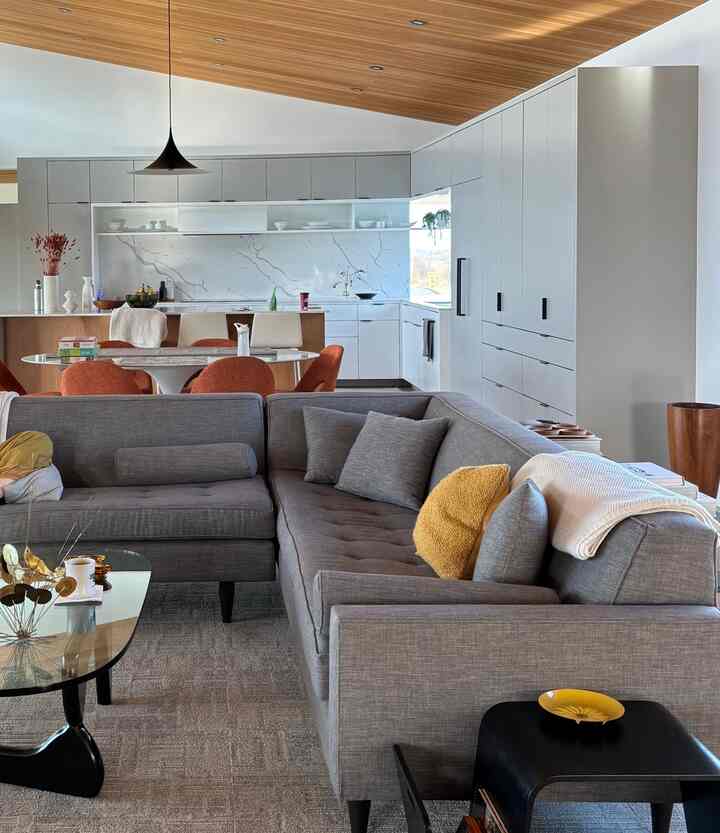 Mid-century modern living room and kitchen with wood tone ceiling, featuring a large gray sectional sofa and orange dining chairs in a stylish open space