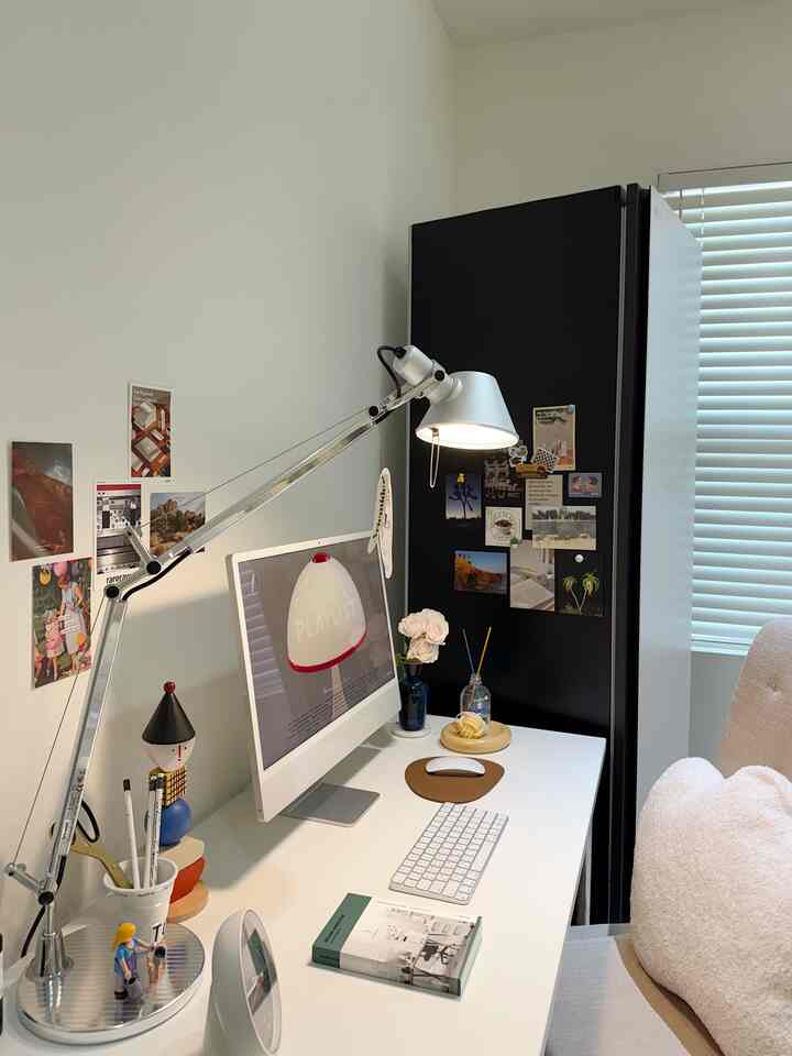 A white-toned compact home office featuring a modern iMac, desk lamp, and comfortable chair in a neat workspace