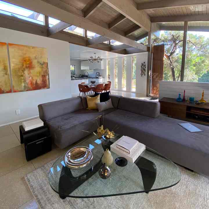 Bright mid-century modern living room with white walls and brown wooden ceiling beams, featuring gray sectional sofa and glass coffee table