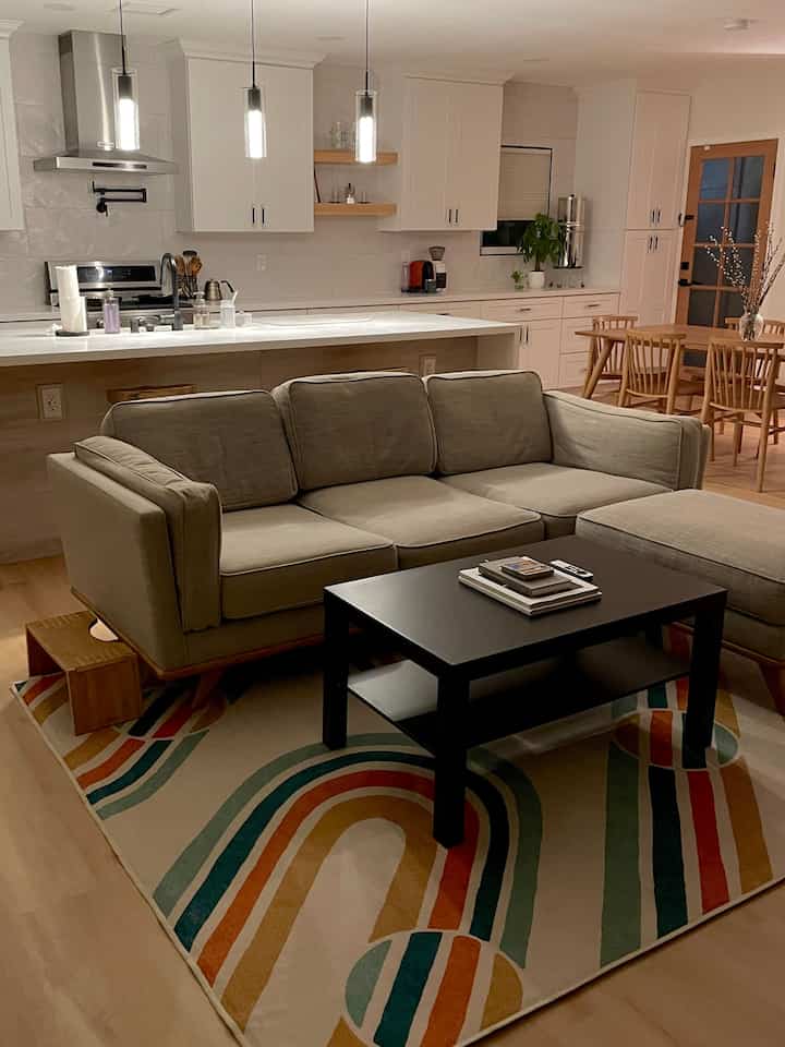 Natural wood tones and white blend in an open living room and kitchen, featuring a beige sofa and colorful rug in a mid-century modern style interior.