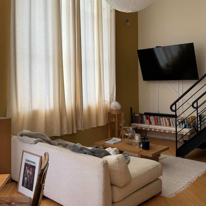 A beige and khaki toned loft living room featuring a cream sofa, wooden coffee table, and bookshelf in a simple natural setting