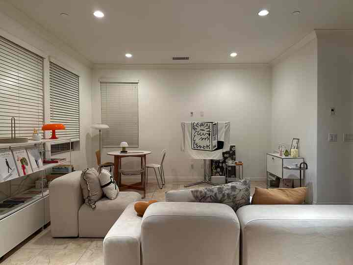 White-toned living room featuring a minimal sectional sofa, round dining table, and table lamps in a simple modern setting