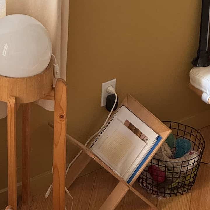 Cozy corner storage space featuring warm natural color walls, wood tone flooring, a wooden stool with table lamp, and a wire storage basket