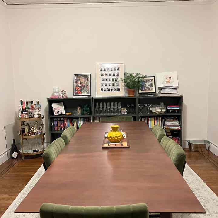 Natural-toned dining room featuring a brown wooden table and green velvet dining chairs, with bookshelves and wall art arranged on the back wall