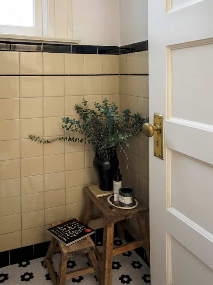 Yellow tiled bathroom corner with wooden stools, a black vase, and a book creating a natural and cozy atmosphere