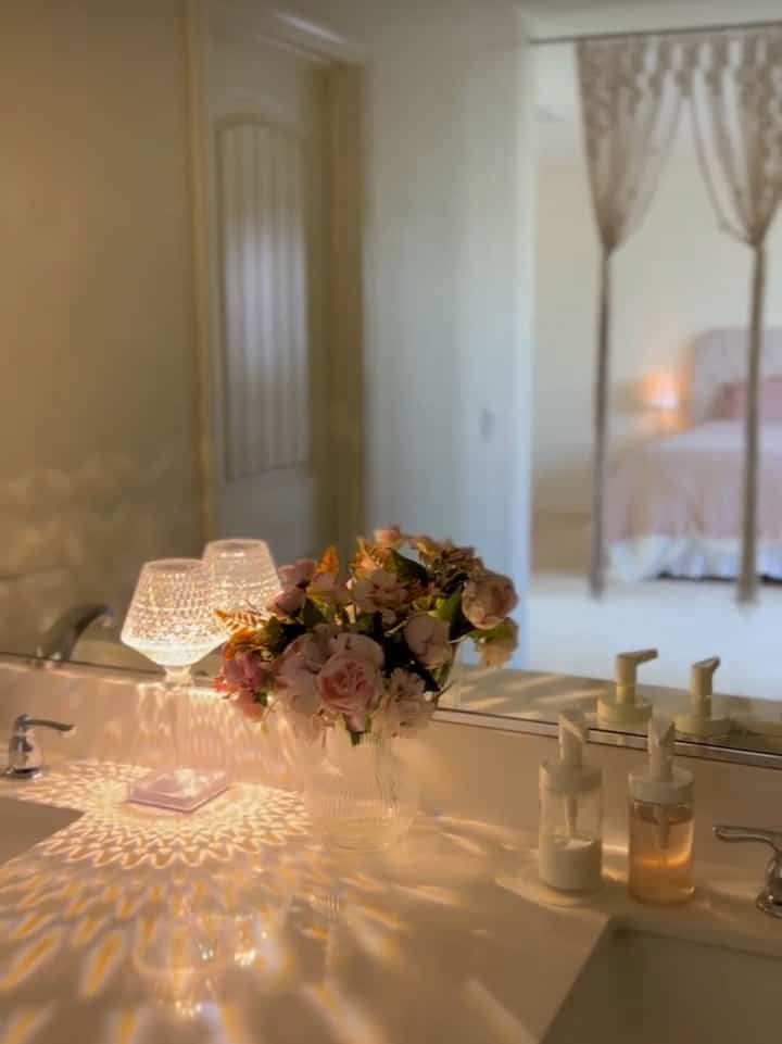 White-toned bathroom with soft cozy lighting, minimal decor including a flower vase and table lamps creating a warm atmosphere
