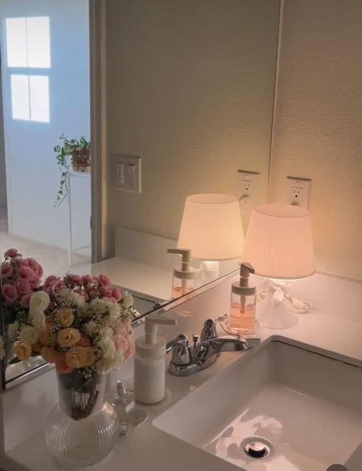 White-toned bathroom basin area featuring warm table lamp and artificial flowers creating cozy mood