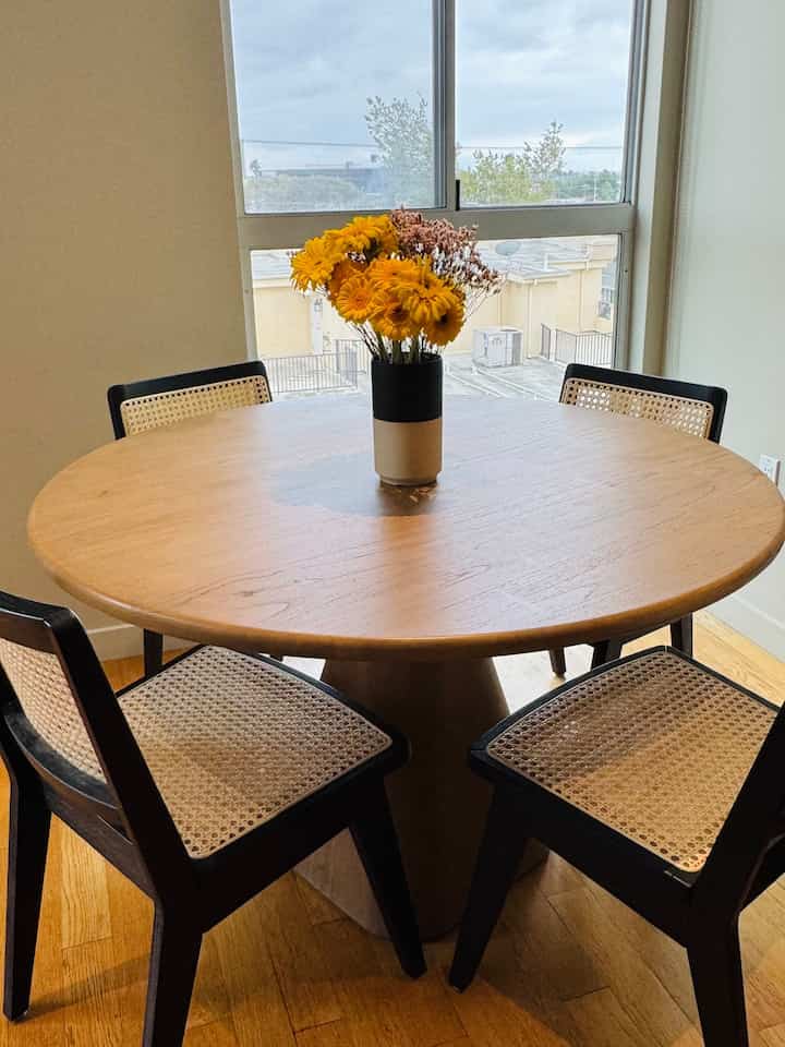 Natural wood tone apartment dining room featuring a round dining table with four woven chairs and a vase of yellow flowers, creating a cozy atmosphere