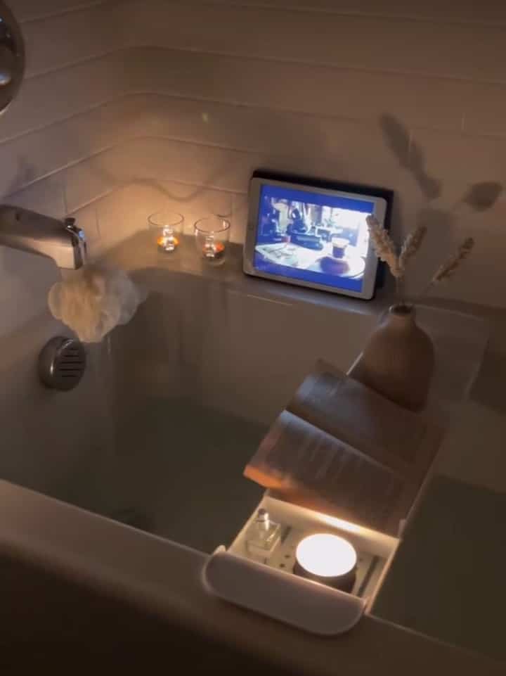 Warm white tone bathroom featuring a bathtub with candles, an open book, and a tablet creating a cozy minimal atmosphere