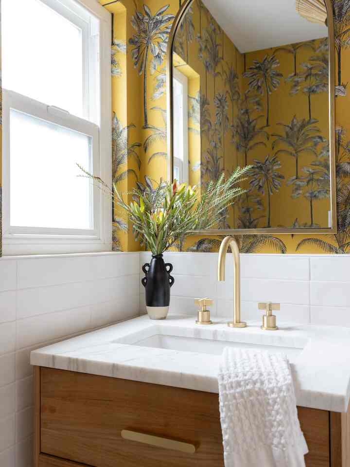 Bathroom with warm atmosphere featuring yellow palm leaf wallpaper, gold faucet, and white marble vanity with mirror