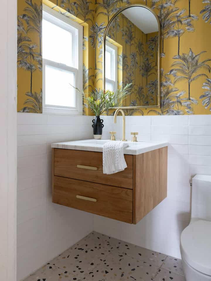 Bright powder room with yellow wallpaper and natural wood vanity featuring gold faucet and arched mirror