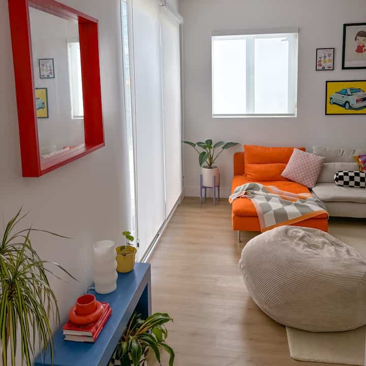 A cozy living room with white walls and wood tone flooring featuring an orange sofa, large bean bag, blue console table, and plants