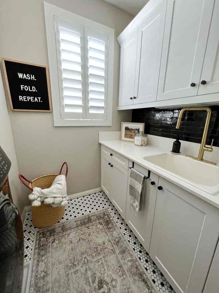 Bright white and beige laundry room featuring white cabinets, gold faucet, a patterned rug, and woven basket for a clean organized look