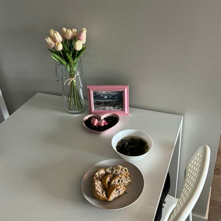 A simple modern dining space for two with beige walls, white dining table and chair, featuring artificial tulips and a pink picture frame