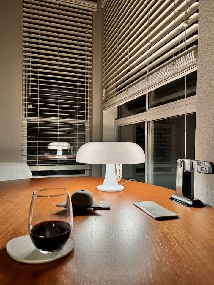 Cozy night mood dining space with warm wood tone table, white table lamp, wine glass, and window blinds