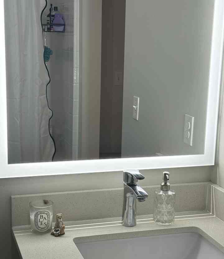 Bright white-toned bathroom space featuring a sink with illuminated mirror, diptyque candle, and transparent soap dispenser in a minimal interior