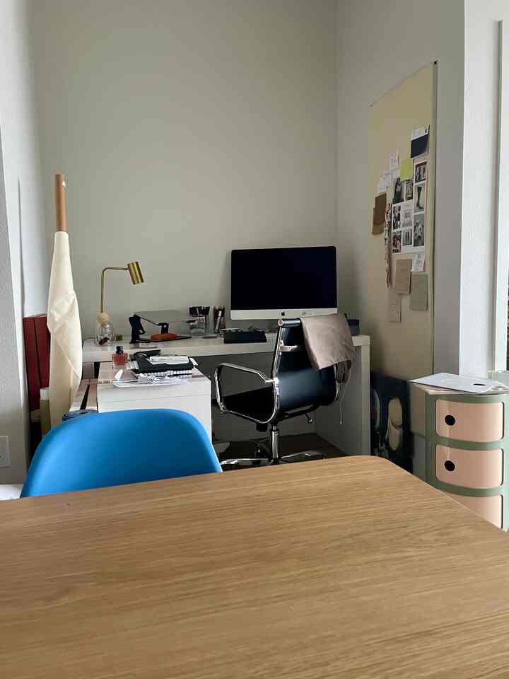 Bright white compact home office featuring a desk with iMac and black office chair, with a wood tone dining table in the foreground