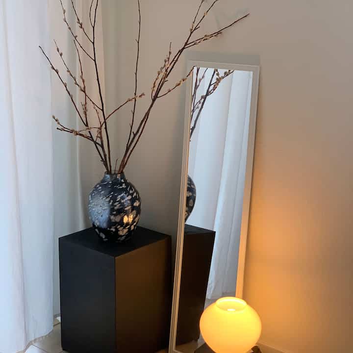 Light gray wall and white curtains background featuring a tall mirror, black metal plinth with a decorative vase and warm glowing lamp in a natural style corner