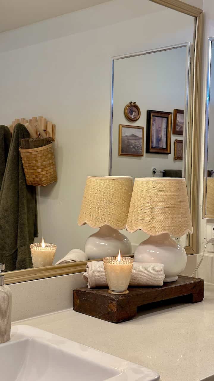 Warm beige and natural toned bathroom featuring a wood tray with candle, table lamps, and towel on countertop, creating a cozy charming space