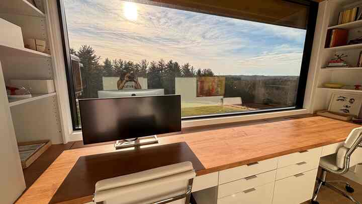 A white and wood tone home office featuring a large desk and panoramic window, flooded with natural light and offering a nature view