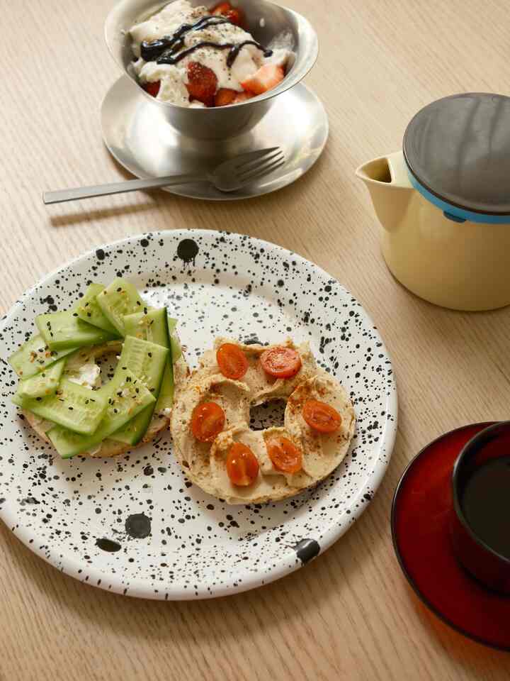 Bright brown wood tone dining table featuring a white splatter-patterned plate with fresh bagels, a bowl of cream and strawberries, coffee cup and teapot in a clean dining room brunch scene