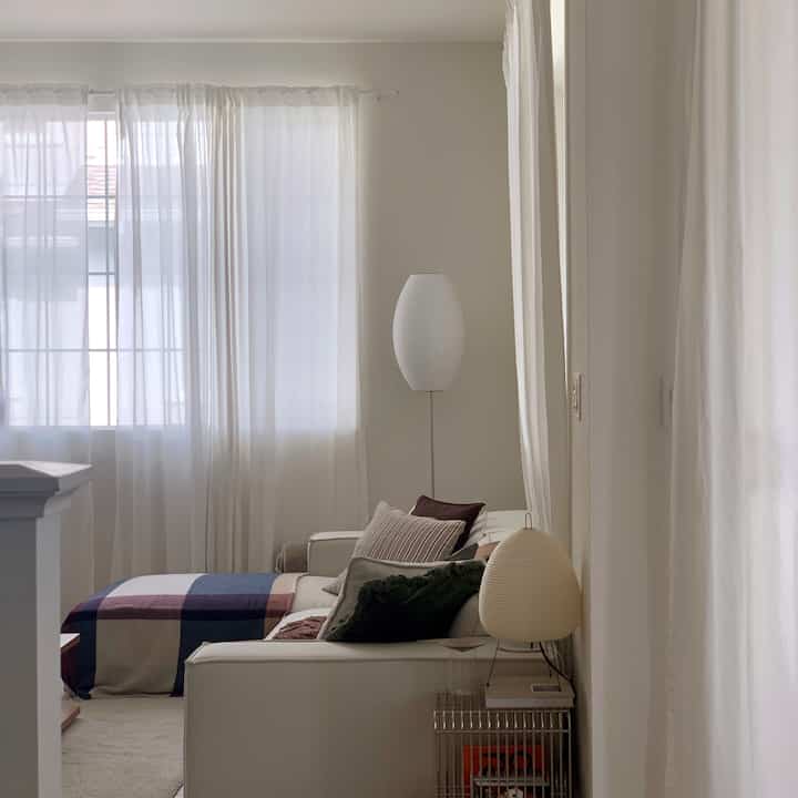 White and natural-toned living room featuring simple sofa, lounge chair, sheer curtains with soft lighting creating serene mood