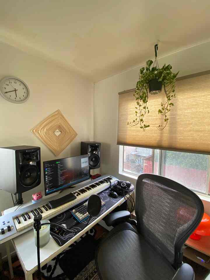 White-walled compact home office featuring a Herman Miller chair, music production keyboard, and monitor speakers with beige blinds