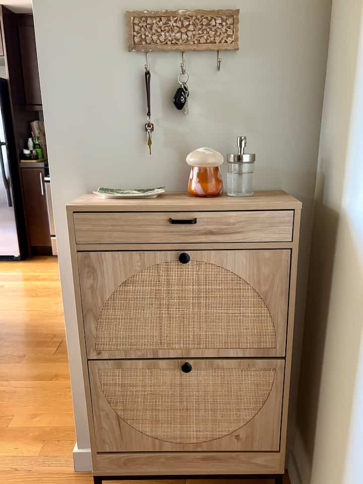 Natural-toned entrance featuring a rattan-accented shoe cabinet topped with hand sanitizer and diffuser, creating a cozy atmosphere