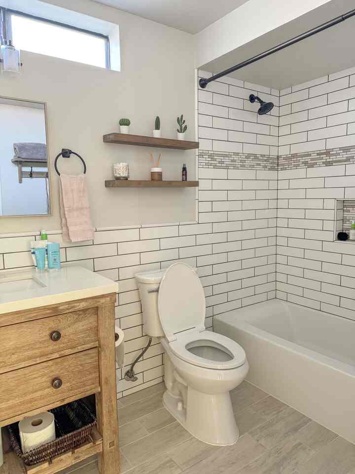 Bright white tiled bathroom featuring natural wood tone vanity and floating shelves, clean shower area with warm natural style