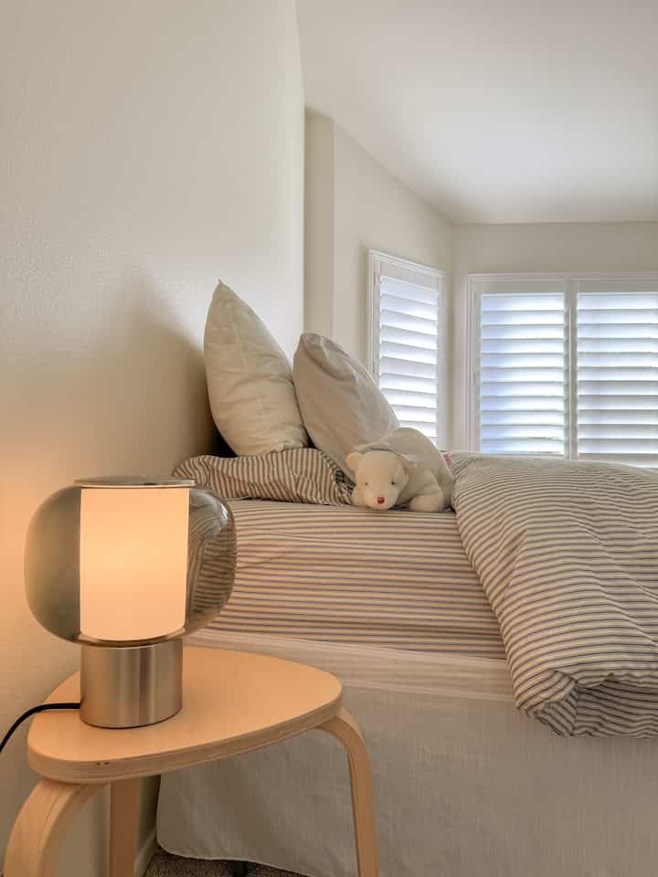 White and blue striped bedding in a bedroom featuring a minimalist wood-tone stool and warm glowing table lamp creating a cozy atmosphere