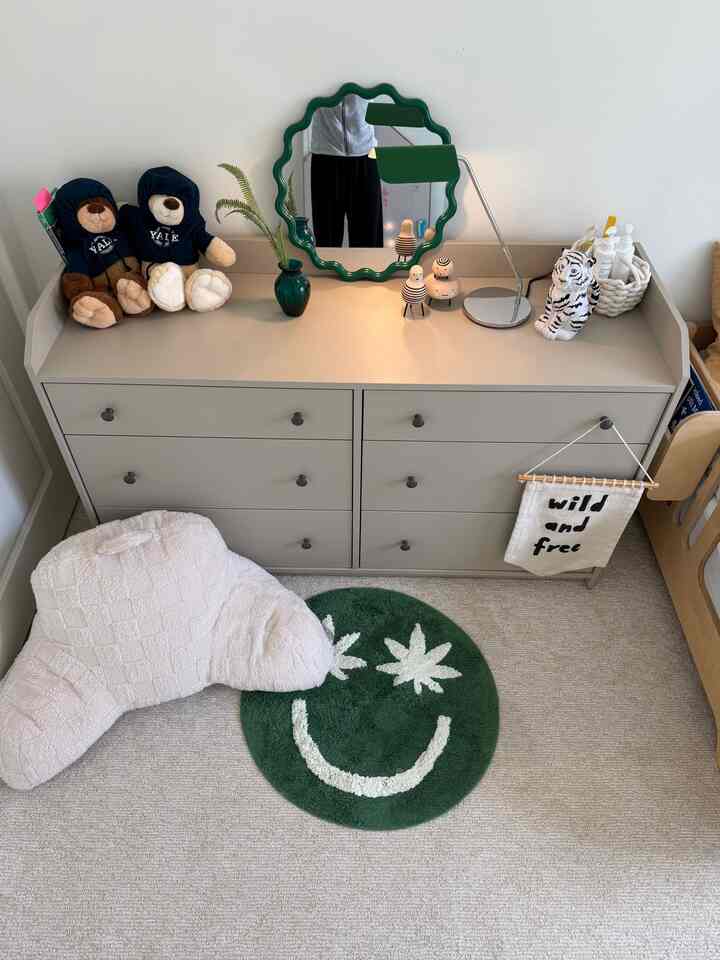 Green and beige toned kids' room featuring a six-drawer dresser with a wavy mirror, table lamp, and children's toys in a minimalist style