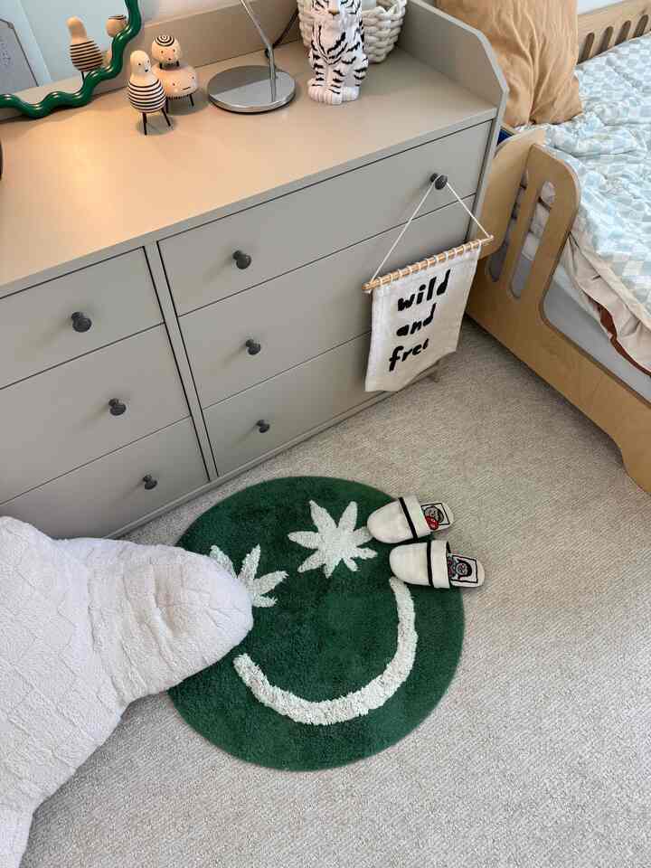 Beige carpeted kids' room featuring green round bath mat, gray dresser, and wooden kids' bed with cozy atmosphere