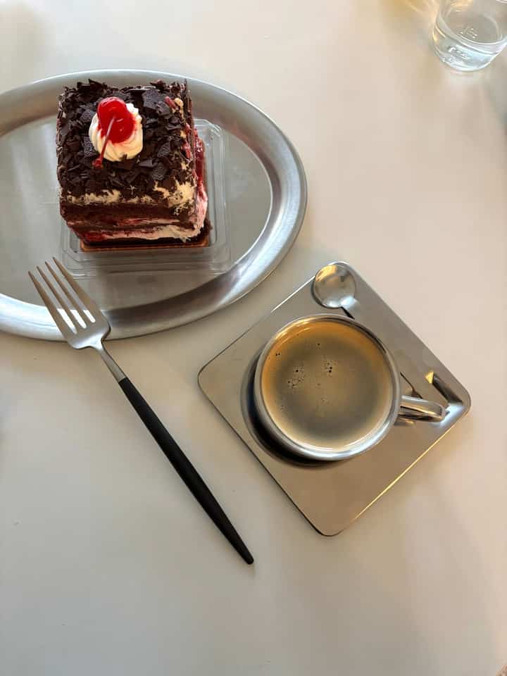 Minimal home cafe scene on white table featuring stainless steel coffee cup, fork, metal trays with a slice of cake