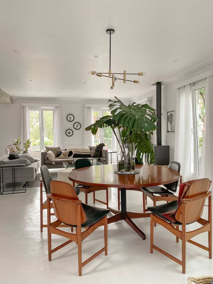 A clean and natural mid-century modern living-dining room in white and wood tones, featuring a central dining table with plant and surrounding sofas