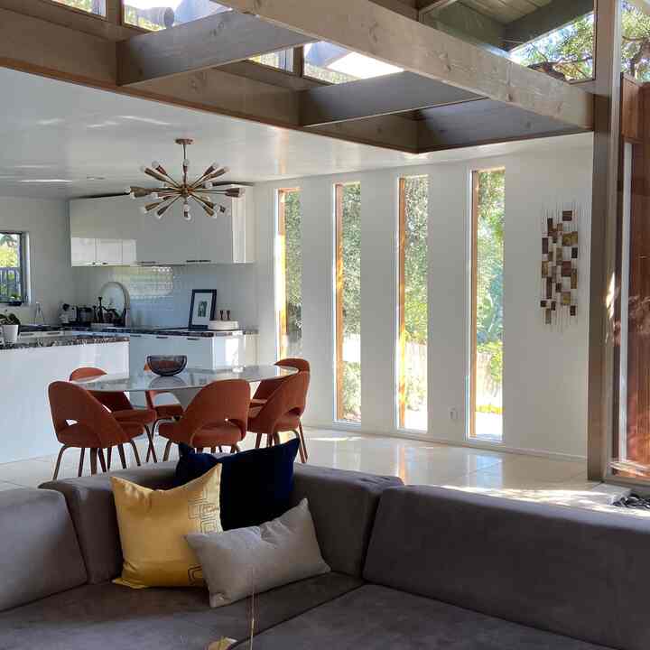 Bright white-walled living and dining area flooded with natural light, featuring orange dining chairs and a gray sofa in Mid-Century Modern style